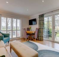 Modern living room with a beige sofa, green and teal armchairs, a round wooden table with purple chairs, and a TV on the wall. Glass doors open to a patio with outdoor seating.