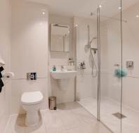 Modern bathroom with beige tiles, featuring a wall-mounted toilet, sink with a mirror, walk-in shower with glass partition, and a towel rack on the left.