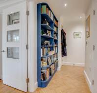 Hallway with blue curved bookshelf filled with books, adjacent to a white painted door and herringbone wood floor. Coats hang in the corner. A green room is partially visible through the open door.
