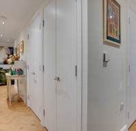 Hallway with wooden herringbone floor, white walls, and multiple doors. Artwork on walls and a small table with decor items in the corner.