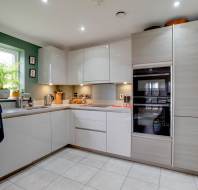 Modern kitchen with white cabinets, stainless steel appliances, and a corner window with plants. A double oven is built into wooden cabinets. The floor is tiled in light gray.
