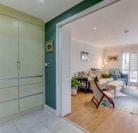 Interior view of a living room with green walls, wooden flooring, sofa, armchair, coffee table, and large windows with shutters, seen through an open doorway with light green cabinets on the left.