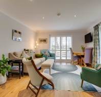 Spacious living room with modern furniture, green and beige chairs, a sofa, round dining table with chairs, large windows with curtains, and wooden flooring. Indoor plants and a wall-mounted TV.
