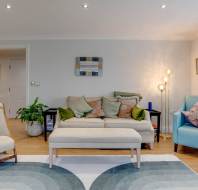 Modern living room with a beige sofa, green and pink cushions, light blue armchair, wooden chair, potted plant, abstract rug, and a floor lamp. Wall mirror and framed art are displayed.