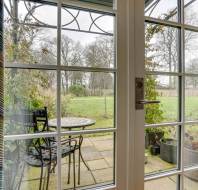 Glass door opens to a patio with a small round table and two chairs. The garden features green grass, potted plants, and trees in the background.