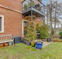 Garden patio with potted plants, a wooden bench, and a small table set. Brick building with a balcony above. Grass lawn and trees in the background.