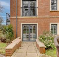 Pathway leads to a modern brick building with large windows and patio doors, surrounded by shrubs and a wooden bench in the foreground.