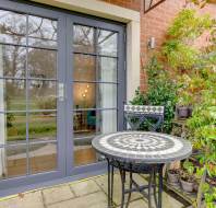 A patio with a round mosaic table and a chair is set in front of glass double doors, surrounded by green plants and shrubs.