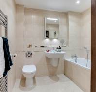 Modern bathroom with beige tiles featuring a wall-mounted sink, mirror, toilet, and bathtub. A heated towel rail with towels is on the left wall.