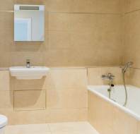 Bathroom with beige tiled walls, a wall-mounted sink below a mirror, a bathtub with a showerhead, and a toilet to the left.