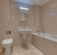 A beige-tiled bathroom with a wall-mounted toilet, a sink with a mirror above, a towel holder, and a bathtub with a hand-held shower.
