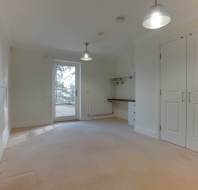 An empty room with light-colored walls and carpet, built-in desk, and double doors. A glass door leads to an outdoor area, and two pendant lights hang from the ceiling.