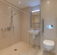 Accessible bathroom with a walk-in shower, wall-mounted sink, and toilet with grab bars. Beige tiles cover the walls and floor.