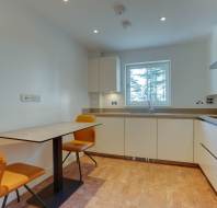 A modern kitchen with white cabinets, a marble-top table, two orange chairs, and a large window.