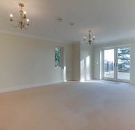 Empty living room with beige carpet, cream walls, and two chandeliers on the ceiling. Two small windows and large glass doors open to a balcony with a view of trees.