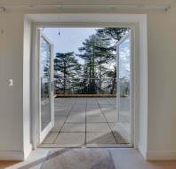 Open double doors leading to a spacious terrace with tile flooring, overlooking trees under a clear blue sky.