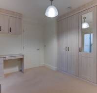 Room with beige carpet, wooden desk with drawers, cabinets above, and large wardrobe with mirrors. Walls are light and two pendant lights hang from the ceiling. Door on the left.