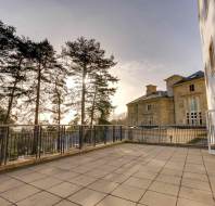Large terrace with stone flooring and railing, adjacent to a historic building. Tall trees in the background with sunlight filtering through.