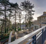 View of a terrace with metal railings, tall trees, and a large building on the right. The sun peeks through the trees, casting light on the scene.