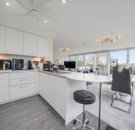 Modern open-plan kitchen and living area with white cabinetry, bar stool, gray dining chairs, large windows, and light fixtures.