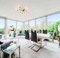Modern living and dining room with glass walls, black and white furniture, and a chandelier. The room opens to a garden balcony with greenery.