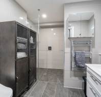 Modern bathroom with a spacious glass-enclosed shower, metal storage cabinet, white sink with mirrored cabinet above, and towel rack. Floor and walls are tiled in light gray tones.