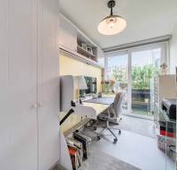 Small home office with a desk, chair, computer, lamp, and shelves. Large window with a view of greenery and a balcony. A storage cabinet is on the right.