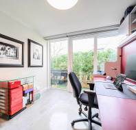 Home office with a desk, chair, large monitor, and wall art. Glass doors reveal greenery outside. Shelves hold various items. Soft natural light fills the room.