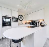 Modern kitchen with white cabinets, a black and white clock, built-in appliances, and a central island with a stove. Ceiling fan and blue sculpture in the background.