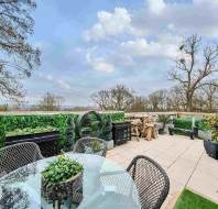 Spacious outdoor terrace with glass table, woven chairs, potted plants, and green hedges, overlooking trees and cloudy sky.
