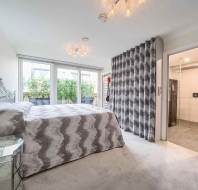 Bedroom with a double bed, gray geometric bedspread, mirrored nightstand, large windows, and an open doorway leading to a bathroom.