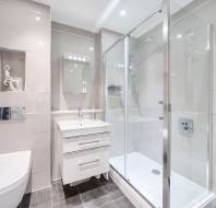 Modern bathroom with a glass shower enclosure, white sink with cabinet, and a wall-mounted toilet. Large mirrors and gray tiled walls create a sleek, spacious look.