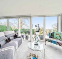 A modern living room with large windows, a gray sectional sofa, colorful upholstered chair, glass coffee table, and abstract sculpture. Outside, potted plants and trees are visible.