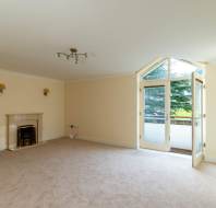Empty living room with beige walls and carpet, featuring a fireplace, ceiling light fixture, and French doors leading to a small balcony with a view of trees.