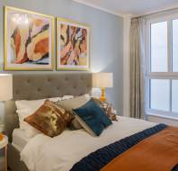 Modern bedroom with a gray upholstered bed, colorful pillows, and two abstract paintings above. Room features large window, white curtains, and bedside tables with lamps.