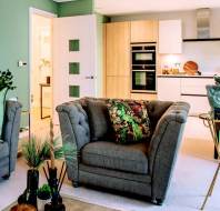 Modern living room with green walls and a beige open kitchen. Features a gray tufted sofa with floral cushions, a wooden coffee table, and decorative plants. Doorway leads to a lit interior hallway.