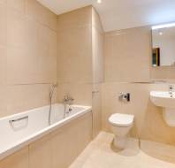 Modern bathroom with beige tiled walls and floor, featuring a bathtub, wall-mounted toilet, sink, and a rectangular mirror above the sink.