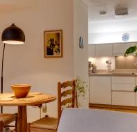 A cozy dining area with a round wooden table and two chairs, adjacent to a modern kitchen with white cabinets. A floor lamp and potted plant add warmth to the space.
