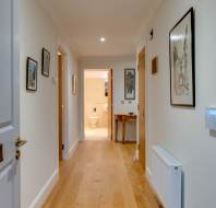 A hallway with wooden flooring leads to a bathroom. The walls are decorated with framed artwork, and a keypad is mounted on the right wall near a radiator.