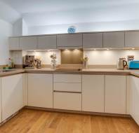 Modern kitchen with beige cabinets, wooden floor, and stainless steel appliances. Countertop holds a coffee machine, kettle, toaster, and utensils. Clock on wall shows 10:10.