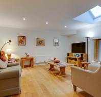 Bright living room with wooden flooring, a skylight, a beige sofa, armchairs, a TV, and framed art on the walls. Coffee and side tables are placed on a rug.