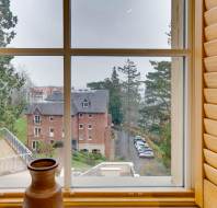 View from a window with wooden shutters showing a red brick building, parked cars, and trees under an overcast sky. A pottery vase is on the window sill.
