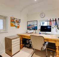 Home office with a wooden desk, computer, filing cabinet, corkboard, medals on the wall, a clock above the window, and a bicycle partially visible on the left.