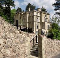 Large stone mansion with tall columns and ornate details, surrounded by lush greenery. Stone wall and stairs lead up to the entrance. Bright, partly cloudy sky in the background.