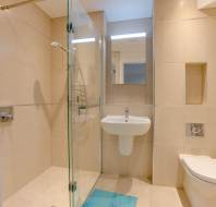 Modern bathroom with beige tiles, featuring a glass-enclosed shower, wall-mounted sink, and toilet. A small blue rug is on the floor.