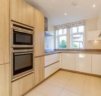 Modern kitchen with light wood cabinetry, stainless steel appliances, and white countertops. A window with floral curtains overlooks a tiled floor.