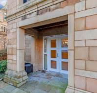 Stone building entrance with a white door and glass panels. Surrounded by trimmed hedges and paving stones.