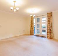 Empty living room with beige carpet, cream walls, and floral curtains, featuring two ceiling lights and a glass door leading outside.