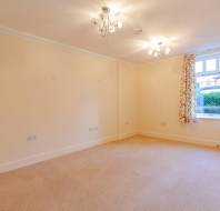 Empty room with beige carpet, cream walls, floral curtains, and two ceiling light fixtures. A single window overlooks greenery outside.