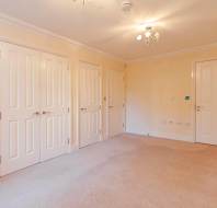 Spacious room with beige carpet, cream walls, three white doors on one side, and a single door on the opposite side, illuminated by a ceiling light fixture.